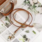 Bandoulière Cuir Fine marron aux coutures visibles avec mousquetons dorés posée près d’un sac classique