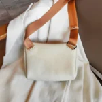Sac à Main Banane Cuir Femme de couleur blanche avec une bandoulière en tissu orange posé sur un vêtement blanc