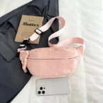 Sac Banane Velours Lisse rose avec sangle ajustable et matière douce Sac Banane Velours Lisse de couleur rose avec une matière douce et une sangle ajustable posé sur une surface blanche à côté d'un livre et d'un smartphone