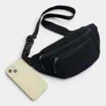 Sac banane velours côtelé noir posé à plat de face, avec une sangle noire à boucle plastique et un smartphone beige