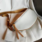 Sac banane cuir grainé blanc posé à plat, bordures caramel et sangle marron avec zip métallique