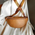 Sac banane cuir grainé camel posé de face sur une veste, avec sangle bicolore et fermeture métallique chromée