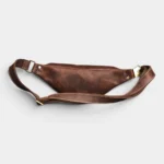 Petit sac banane cuir marron à l’aspect vintage posé à plat vue arrière avec sangle et boucle en métal doré