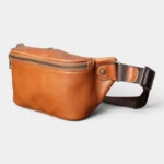 Sac Banane Cuir Vintage Homme Sac banane cuir marron clair posé de trois quarts, avec fermeture métallique sur le dessus et sangle marron foncé
