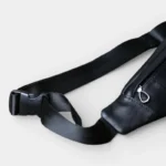 Petit sac banane cuir noir posé de côté, avec sangle réglable noire et boucle clip en plastique