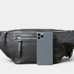 Grand sac banane cuir noir posé de face, avec un smartphone glissé devant la poche zippée