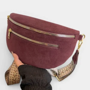 Sac banane en simili cuir suédé bordeaux tenu à la main de face, avec sangle à motif chevron beige et attaches dorées