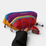 Sac banane multicolore outdoor de face, tenu à la main, avec élastiques violets sur la poche avant rouge