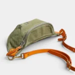 Sac Banane Toile Kaki Militaire Bandoulière Cuir Véritable Sac banane toile kaki militaire posé de côté, avec bandoulière en cuir cognac et mousquetons dorés