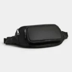 Sac Banane Homme Cuir Grainé Minimaliste Sac banane en cuir grainé noir posé à plat, vue de trois quarts avec poche frontale zippée aux fermetures argentées