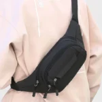 Sac banane en nylon noir porté en bandoulière croisée de face par une personne au style streetwear décontracté