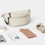 Sac banane nylon froissé beige posé de face, entouré d'accessoires du quotidien dont un smartphone et un portefeuille
