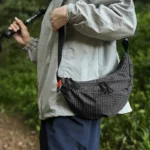 Grand Sac Banane Nylon Ripstop Quadrillé Imperméable Sac banane nylon ripstop quadrillé noir porté en bandoulière par un randonneur, de profil en forêt