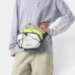 Sac Banane Technique Gorpcore Outdoor Nylon Ripstop Sac banane nylon ripstop jaune et blanc porté à la main par une personne au style gorpcore outdoor, vu de face