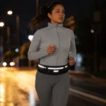 Sac Banane Running Ultra Fin Imperméable Réfléchissant Sac banane running en nylon noir porté à la taille par une femme, de face lors d'une course nocturne en ville