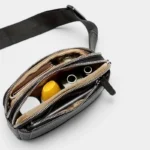 Sac banane en simili cuir noir grainé vu de dessus, poches ouvertes révélant une organisation intérieure avec poches zippées et contenu varié