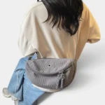 Grand Sac Banane Femme Velours Côtelé Demi-Lune Sac banane en velours côtelé gris porté en bandoulière par une femme de dos, au style décontracté-chic