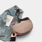 Grand Sac Banane Femme Velours Côtelé Demi-Lune Sac banane en velours côtelé taupe tenu à la main, présenté de face sur une veste en jean