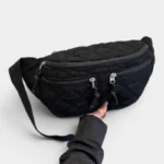 Grand Sac Banane Femme Velours Côtelé Matelassé Grand sac banane en velours côtelé matelassé noir, posé de face avec une main tenant la sangle