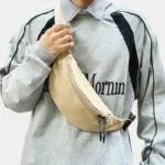 Sac Banane Nylon Satiné Léger Sac banane nylon satiné beige porté en bandoulière sur la poitrine avec sangle tenue à la main