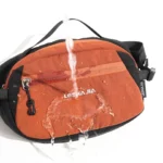 Sac Banane Bicolore Randonnée Nylon Léger Eau versée sur un sac banane bicolore orange et noir, les gouttes perlent à la surface du tissu.