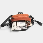 Sac Banane Bicolore Randonnée Nylon Léger Sac banane bicolore orange ouvert vu du dessus, contenant des lunettes de soleil, un carnet et un téléphone.
