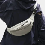 Sac Banane Nylon Satiné Léger Sac banane nylon satiné gris clair porté en bandoulière à la hanche avec boucle clip noire