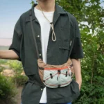Sac Banane Randonnée Ultraléger Double Sangle Sac banane beige et vert porté en bandoulière par un homme en randonnée avec seconde sangle bouclée à la taille