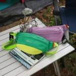 Sac Banane Coupe-Vent Années 90 Déperlant Deux sacs banane coupe-vent années 90, vert émeraude et violet lilas, posés sur une table de camping