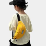 Sac banane enfant en toile de nylon jaune porté en bandoulière dans le dos par un petit garçon vu de trois quarts