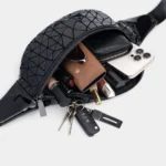 Sac banane en PVC noir brillant, poche principale ouverte laissant apparaître des cosmétiques et des clés