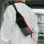 Sac Banane Rectangulaire Allongé Nylon Ripstop Déperlant Sac banane en nylon ripstop noir porté en bandoulière dans le dos par un homme, fermeture rouge apparente.