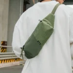 Sac Banane Rectangulaire Allongé Nylon Ripstop Déperlant Sac banane en nylon ripstop vert kaki porté en bandoulière dans le dos par un homme, sangle large sur l'épaule.