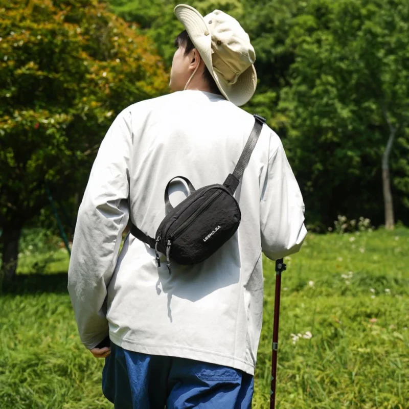 Sac banane en nylon froissé noir porté dans le dos par un homme en randonnée dans une prairie boisée