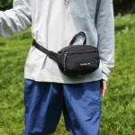 Sac banane en nylon froissé noir porté à la taille de face par un homme dans une prairie