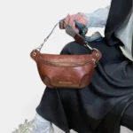 Sac banane simili cuir cognac tenu à la main par une femme au style chic-décontracté, avec une sangle ethnique bleue et dorée