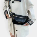 Sac banane simili cuir noir porté en bandoulière par une femme de face au style chic casual avec sangle ethnique dorée