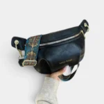 Sac banane en simili cuir noir tenu à la main, de face, avec une sangle ethnique bleue et dorée à mousqueton doré