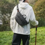 Sac banane noir porté dans le dos par un randonneur tenant un bâton de marche en prairie