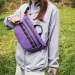 Sac banane violet porté en bandoulière par une randonneuse le soulevant par sa poignée noire