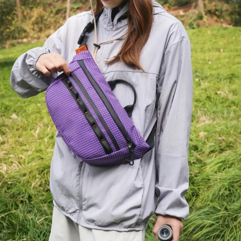 Sac banane violet porté en bandoulière par une randonneuse le soulevant par sa poignée noire