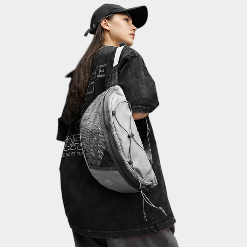 Sac banane gris porté en bandoulière sur le torse par une femme de trois quarts au style streetwear