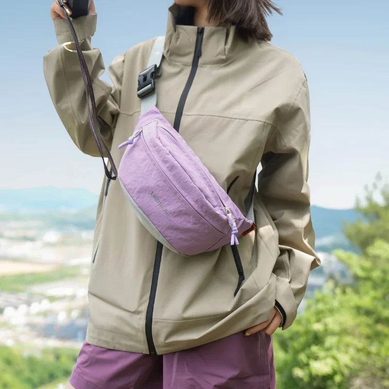 Sac banane coupe-vent années 90 violet lilas porté en bandoulière par une femme lors d'une balade en montagne