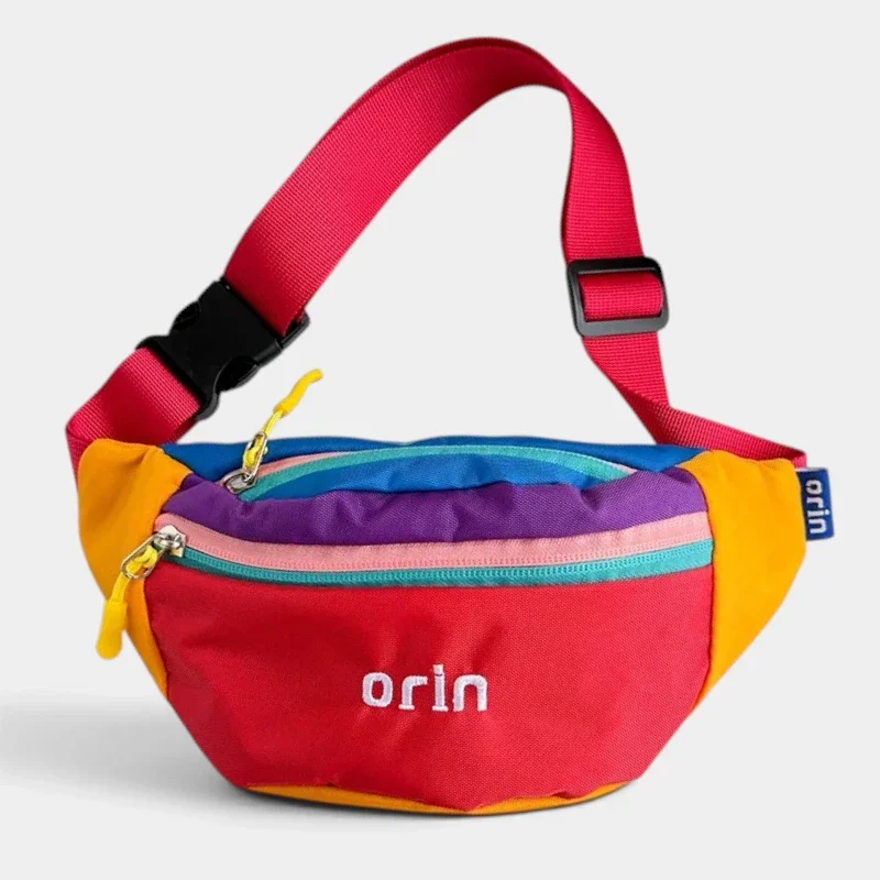 Sac banane enfant en nylon rouge à empiècements colorblock, suspendu par sa sangle et vu de trois-quarts.
